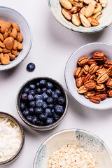 Ingredients for homemade granola: oatmeal, coconut crunch, pecans, almond, brazilan nuts, blueberries in a bowls on light background. Healthy diet breakfast. Flatlay. Selective focus