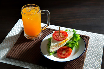 Sandwich cheese and ham and fried egg with veg. in white dish on dark brown apron and oranges juice in glass