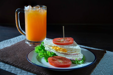 Sandwich cheese and ham and fried egg with veg. in white dish on dark brown apron and oranges juice in glass