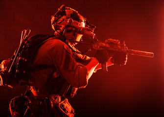 Army special operations soldier, commando fighter in full tactical ammunition, helmet with radio headset and night vision device, aiming short barrel assault rifle in darkness, low key studio shoot