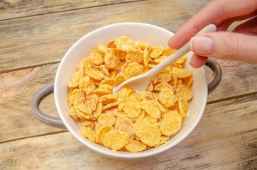girl's hand with a white spoon mixes corn flakes in a bowl with milk on a wooden background