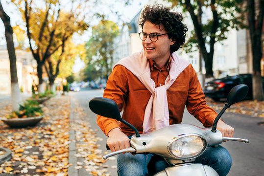 Outdoor image of a handsome young man with curly hair, driving on his scooter, dressed in a casual outfit and transparent eyeglasses, has joyful expression.