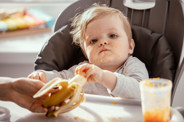  Baby in the child dining chair eat healthy, vegetable food. Child eat  pumpkin and carrot puree with banana. Mother's hand. Dirty face of happy kid. Portrait of a baby eating with a stained face.