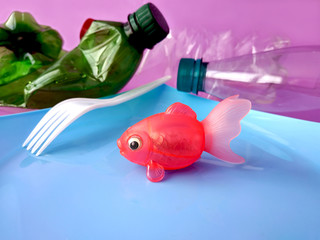 Plastic model of a fish with a plastic fork on a plastic food dish in the concept of environmental pollution against the background of used plastic bottles.