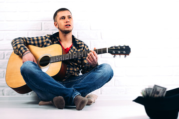 the man playing the guitar sitting against a brick wall at the front of the hat with money