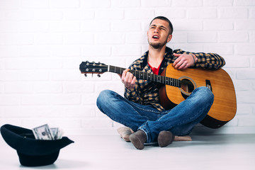 the man playing the guitar sitting against a brick wall at the front of the hat with money