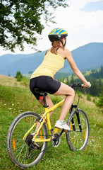 Fototapeta premium Back view of attractive woman cyclist cycling on yellow mountain bicyclev on a grassy hill, wearing helmet, enjoying summer day in the mountains. Outdoor sport activity, lifestyle concept