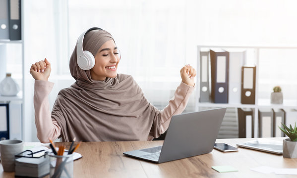 Joyful Arabic Woman In Hijab Listening Music In Headphones At Workplace