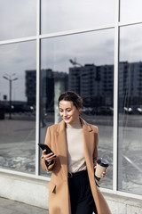 Business Woman smiling charmingly, using her Smartphone. Female enjoying sunny day and funny conversation. Typing on her Phone with Interest. Brunette Girl. Successful Woman. Phones. Apps.