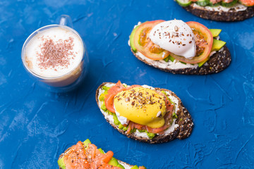 Sandwiches with smoked salmon, eggs, sauce and avocado on blue background. Cup of coffee with cinnamon. Concept of breakfast and healthy nutrition.