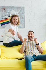 excited homosexual men smiling near lgbt flag in living room