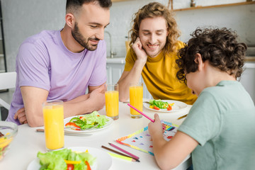 mixed race kid drawing picture with lgbt flag near happy homosexual parents