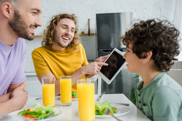 happy homosexual man pointing with finger at digital tablet with blank screen near mixed race son