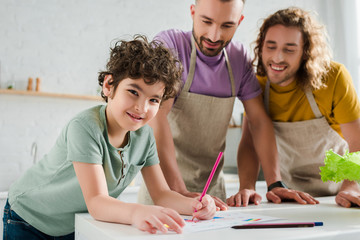 selective focus of happy mixed race kid drawing picture near happy homosexual parents