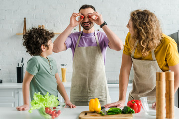 happy and bearded homosexual man covering eyes with cherry tomatoes near mixed race son and partner
