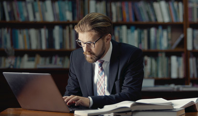 Busy businessman in formal suit and glasses typing on laptop keyboard sitting among books in library