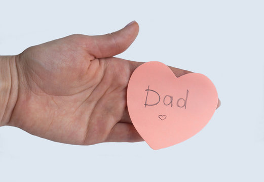 Woman's hand holding pink heart-shaped sticky note with hand written word "Dad" on it on white background. Concept of love, minimalism, Father's Day.