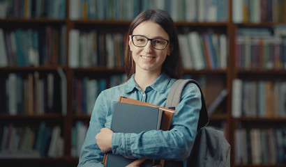 Portrait of lovely smiling female student with glasses and backpack holding books looking camera