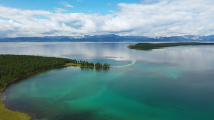 Bird's-eye view of the blue lake Khubsugul in Mongolia, against a background of blue mountains in a haze and the sky with clouds, forests, green grass. Below, white seagulls fly by.