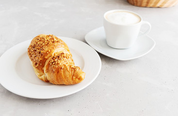 Classic french croissant sprinkled with nuts on a white porcelain plate and a white cup with cappuccino next to it on a gray concrete table. Breakfast in the cafe. Coffee break. Place for text