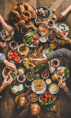 Turkish breakfast. Flat-lay of peoples hands taking and eating Turkish pastries, vegetables, greens, cheeses, fried eggs, jams and tea in copper pot and tulip glasses over wooden background, top view