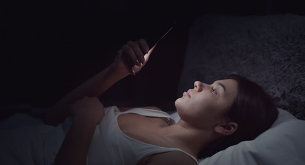 Young woman lying in bed in dark room using smart phone before going to sleep, addiction concept