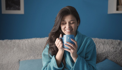 Content charming brunette in blue bathrobe sitting on sofa holding cup with closed eyes in morning