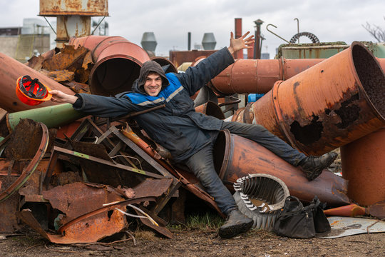 Factory Roof. Work At Height. A Working Man Lies On A Pile Of Metal Pipes And Scrap Metal. In A Hand A Working Helmet. Jokes In The Workplace. Neglect Of Remedies. Safety Violation