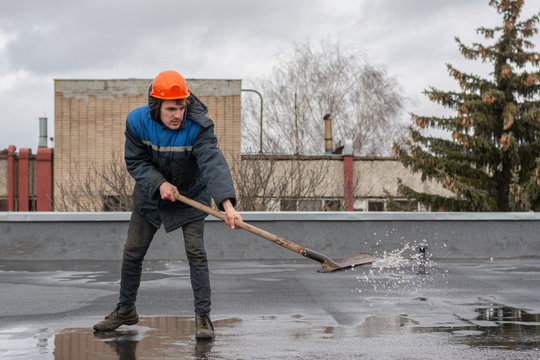 Worker builder on the roof of the factory removes water from the puddle with a shovel. on the head a construction helmet. work at height.