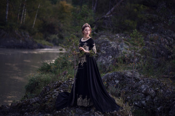 A girl in a black dress with a crown on her head stands on the banks of the river