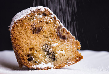 Sprinkling icing sugar powder over the piece of fresh sweet muffin cake with raisins close up on black background.