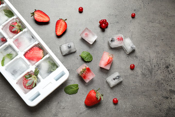 Ice cubes with berries and tray on grey table, flat lay