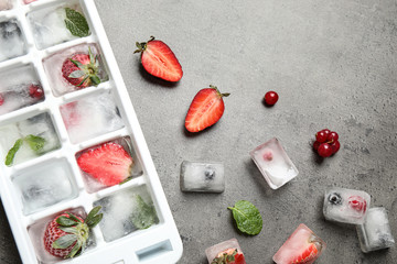 Ice cubes with berries and tray on grey table, flat lay