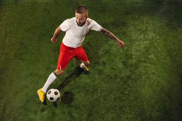 Top view of caucasian football or soccer player on green background of grass. Young male sportive model training, practicing. Kicking ball, attacking, catching. Concept of sport, competition, winning.