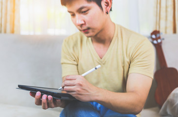 The young man uses a digital tablet with a deer acoustic ukulele on the back of the man. To rest on the weekend, Focus on the hand that is holding the digital pen.