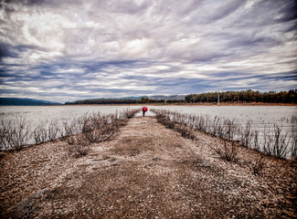 Mujer con paraguas rojo al final del camino de tierra que llega al lago y con nubes de tormenta