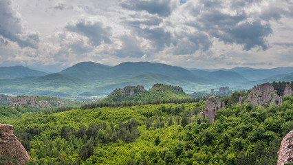 Fototapeta na wymiar Panoramic view of a green mountain valley with dense forests and scattered large rock formations, Belogradchik Rocks, Bulgaria. Mountains and dramatic cloudy sky in the background.