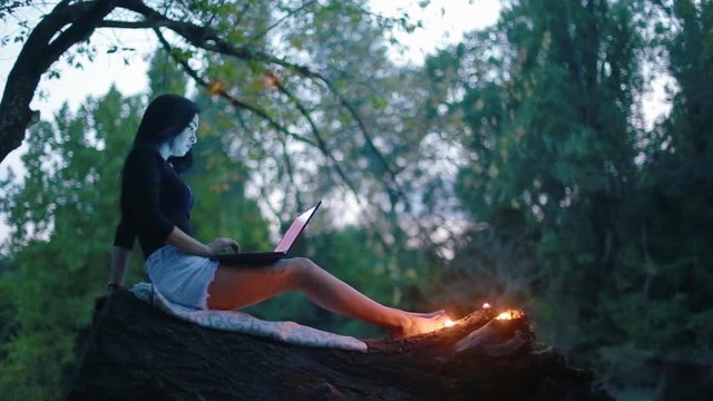 Charming sexy young brunette woman sits on trunk of fallen tree and works on laptop and near her feet burning candles in forest in evening time