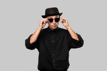 Portrait of a stylish man in a black shirt and sun glasses hat isolated over grey background.