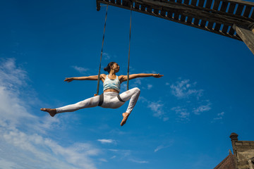 outdoors portrait of young happy and athletic Asian Indonesian woman doing aero yoga workout training body balance  hanging from swing rope isolated on blue sky