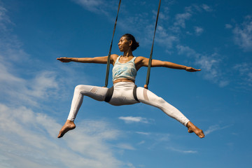 outdoors portrait of young happy and athletic Asian Indonesian woman doing aero yoga workout training body balance  hanging from swing rope isolated on blue sky