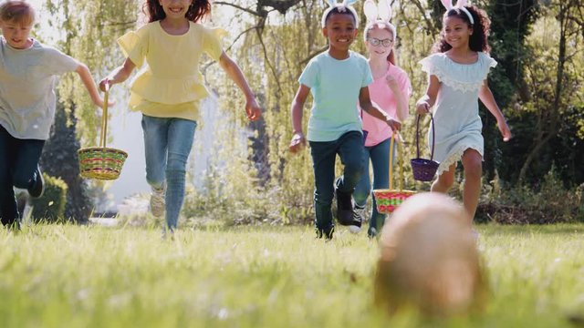 Group Of Children Wearing Bunny Ears Running To Pick Up Chocolate Egg On Easter Egg Hunt In Garden