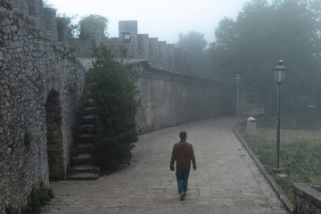 Man walking alone at distance. Fortification on top of the mountain, old castle. Journey. Mystical atmosphere, fog, white haze, mist. Summer day.