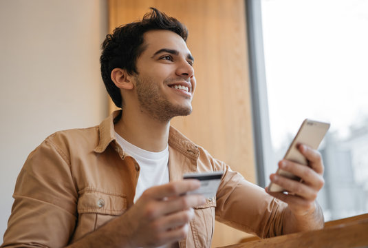 Indian Man Holding Credit Card, Using Smartphone And App For Online Shopping, Mobile Banking. Young Asian Freelancer Receive Payment. Happy Emotional Hipster Ordering Food Online, Booking Tickets