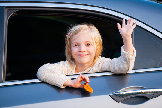 A Little Girl Is Sticking Her Head Out The Car Window. Road Trip Or Travel Concept. Happy Kids Travel By The Car. Cute Child Leaning Out A Van Window, Smiling And Waving.