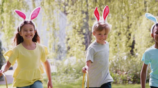 Group Of Children Wearing Bunny Ears Running On Easter Egg Hunt In Garden