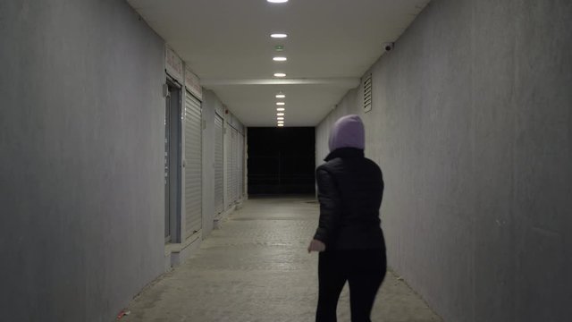Frightened girl in a hoodie is running away from her pursuer through an underground pedestrian crossing. Long dark corridor with blinking light and shops with lowered roller shutters. Crime concept.