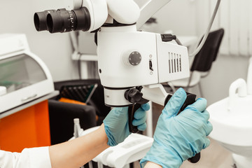 Young woman dentist doctor looks through a professional microscope in a dental clinic. A doctor in a disposable medical mask and cap. Advanced equipment in dentistry