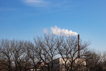 Industrial chimney with smoke and treetops against blue sky