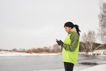 Content attractive female jogger in ear buds standing at riverside and checking fitness app on phone